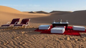 Eine Wüstenlandschaft mit zwei Liegestühlen, einem Tisch mit roter Tischdecke und weißen Kissen im Sand.