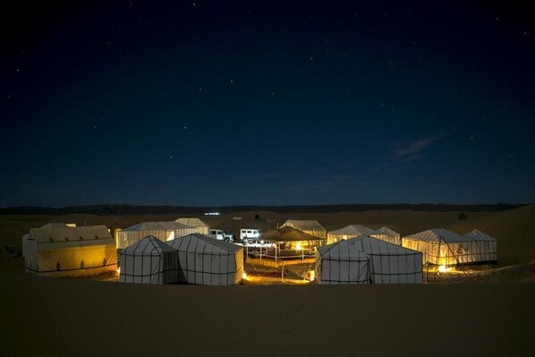 A series of tents illuminated in the desert at night, with a vehicle parked in the center.
