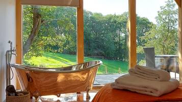 A bathroom with a bathtub, a window with a view of a green landscape, and a bed with an orange blanket.