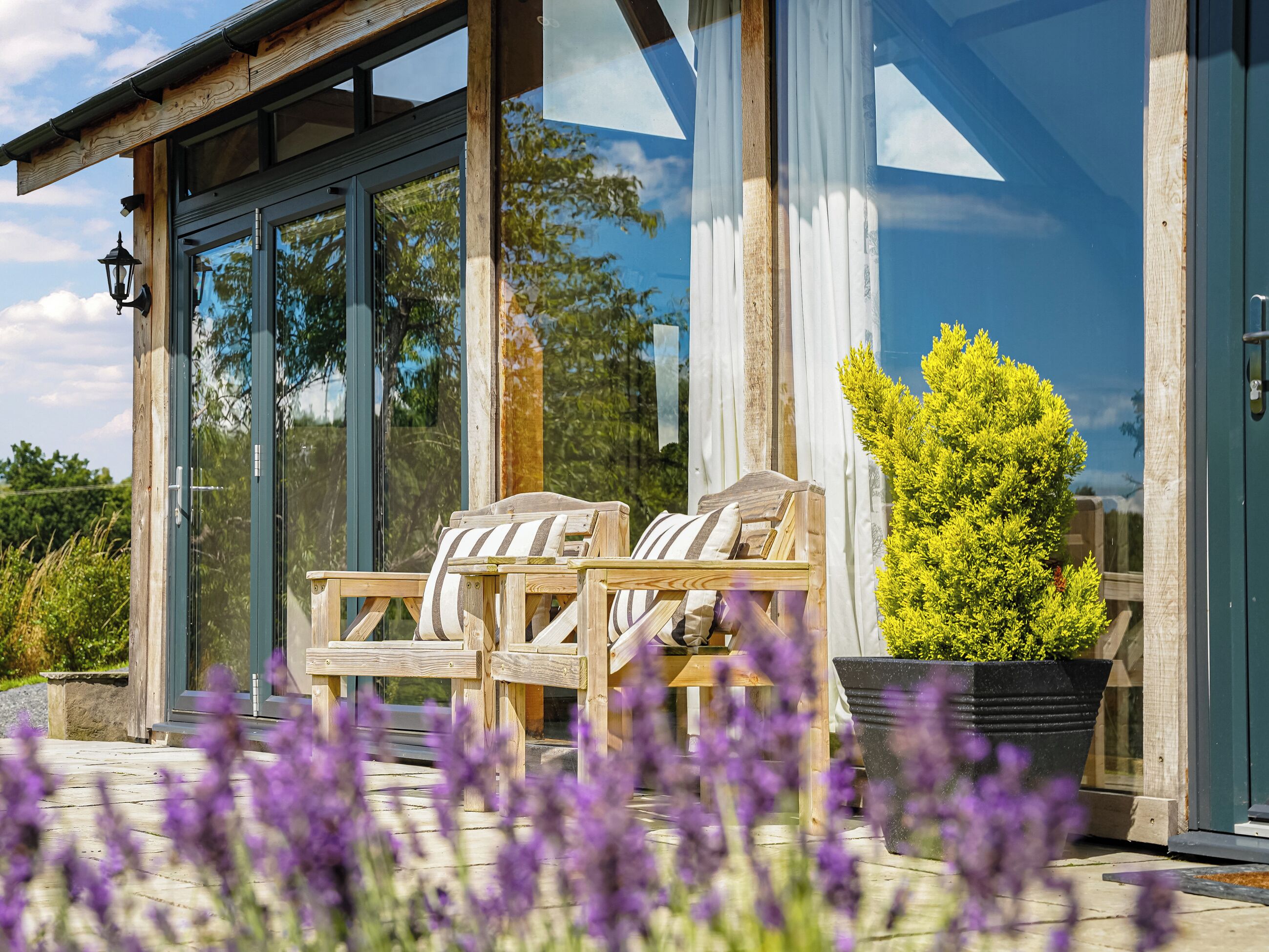 A wooden deck with two chairs and a potted plant, a glass door, and a view of trees and sky.