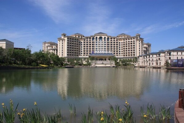 A large, multi-story hotel with a symmetrical facade, a central fountain, and a calm lake in the foreground.