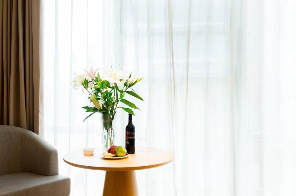 A round wooden table with a vase of white flowers, a bottle of wine, and a plate of fruit.