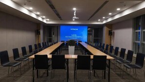Een vergaderruimte met een lange tafel, zwarte stoelen en een groot scherm waarop 'HITACHI LCD MULTIMEDIA PROJECTOR' staat.