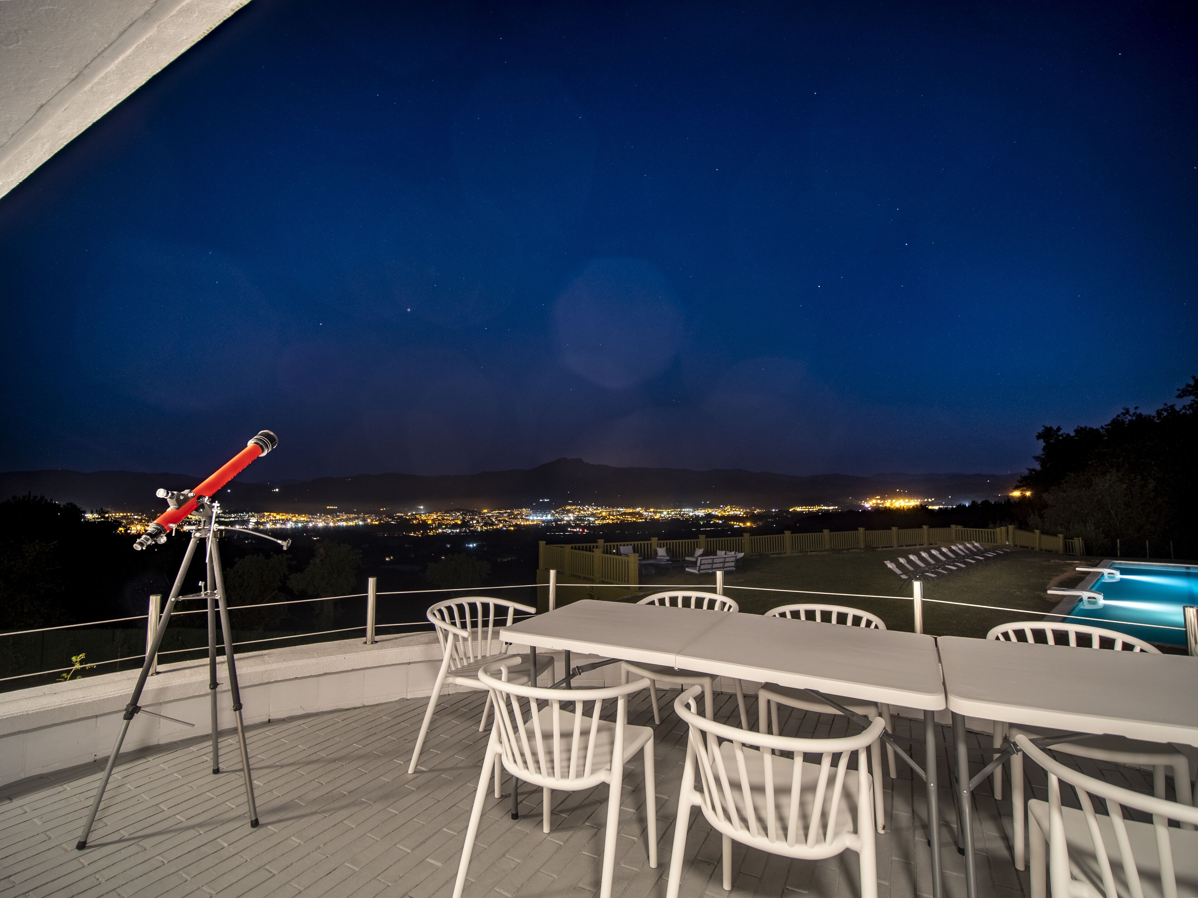 A telescope on a tripod, outdoor dining area with white chairs, and a pool with blue lighting.