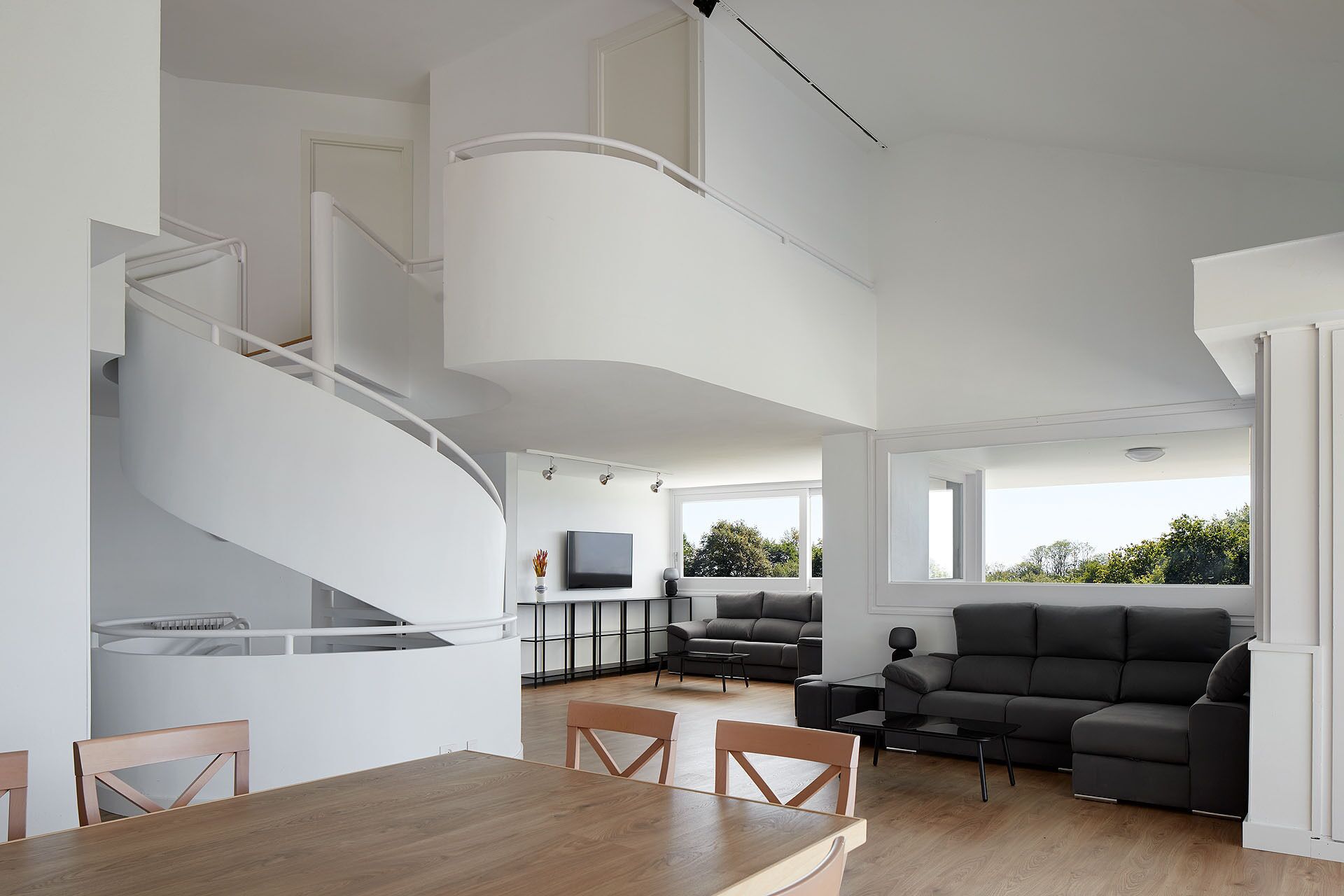 A modern interior with a spiral staircase, a dining area, a living room with a sofa and coffee table, and large windows offering a view of greenery.