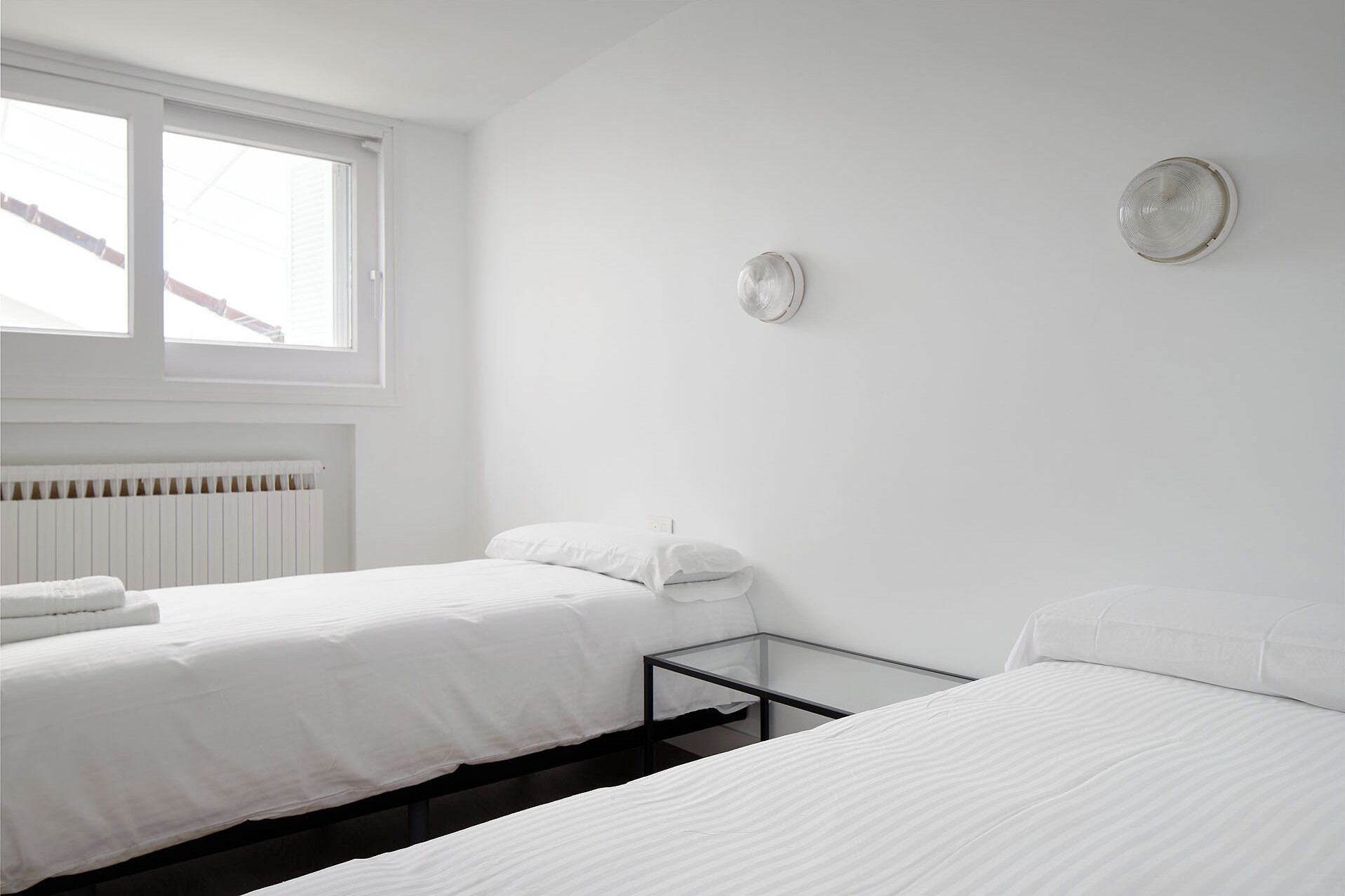 A minimalist bedroom with two beds, a radiator, and a window.