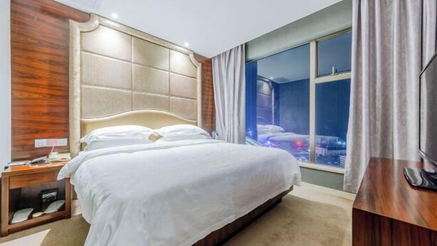 A hotel room with a large bed, a wooden headboard, a nightstand, a TV, and a window with a city view. - Jiuzhou International Hotel (Yangxi)