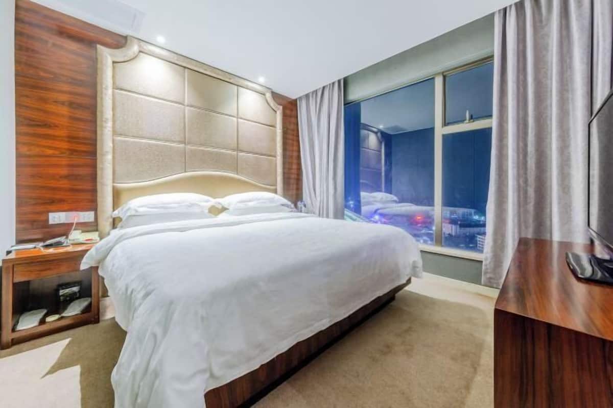 Une chambre d’hôtel avec un grand lit, une tête de lit en bois, une table de chevet, une télévision et une fenêtre donnant sur la ville.