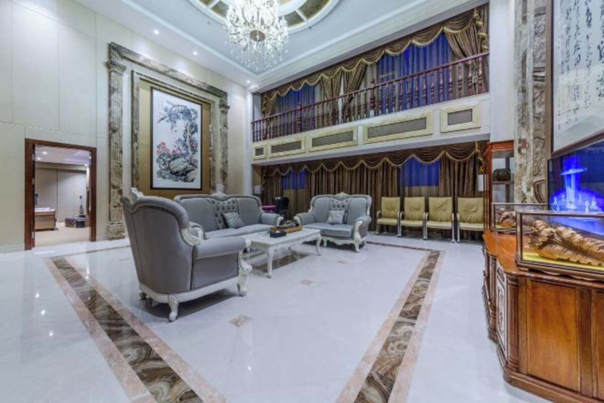 A spacious living room with a high ceiling, a chandelier, a large painting, and a balcony with curtains.