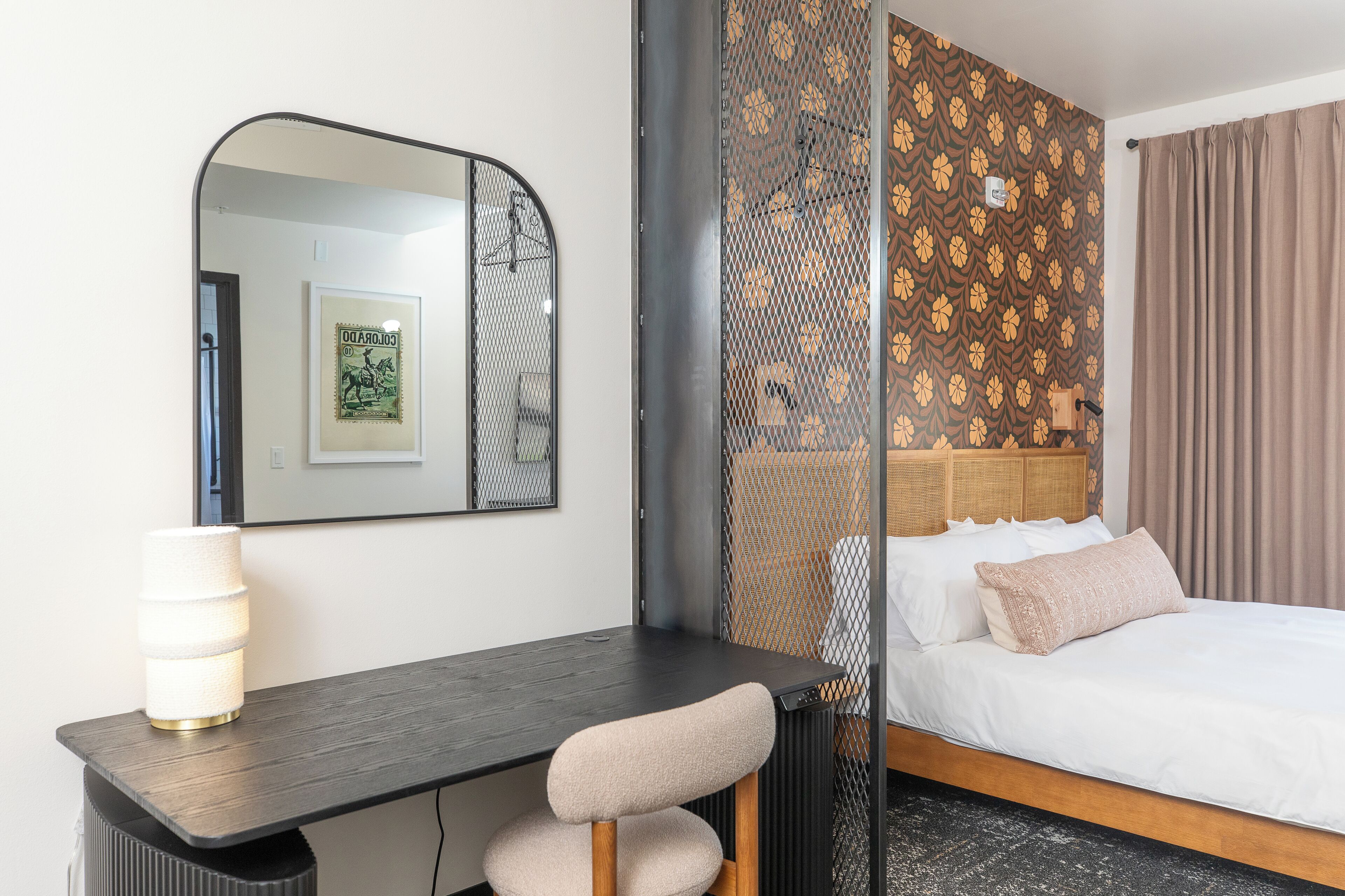 Habitación de hotel con un escritorio de madera oscura, una silla, una cama con ropa blanca, un espejo grande y un panel decorativo en la pared.