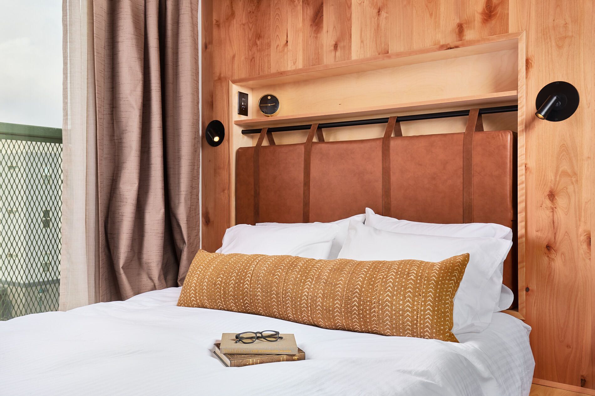 Un lit avec du linge de lit blanc, un oreiller marron à motifs et un livre sur lequel reposent des lunettes.