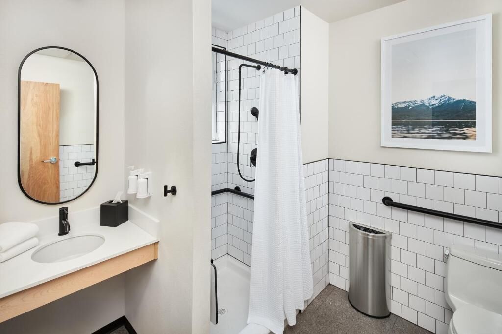 A modern bathroom with a white sink, a mirror, a shower with a white curtain, a toilet, and a framed picture on the wall.