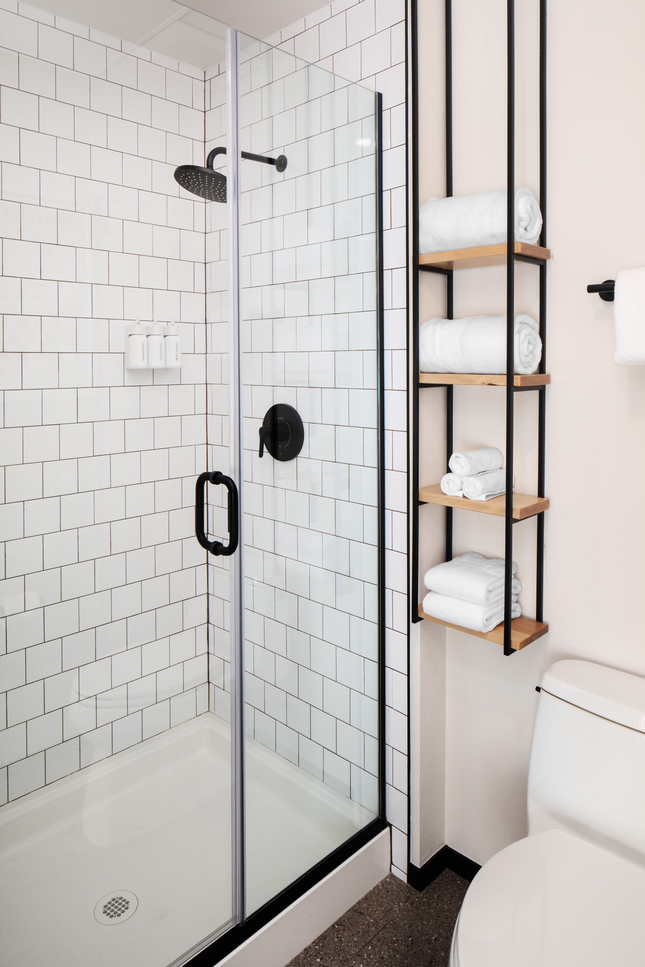 A glass-enclosed shower with white tiled walls, a black door, and a shelf with neatly folded white towels.