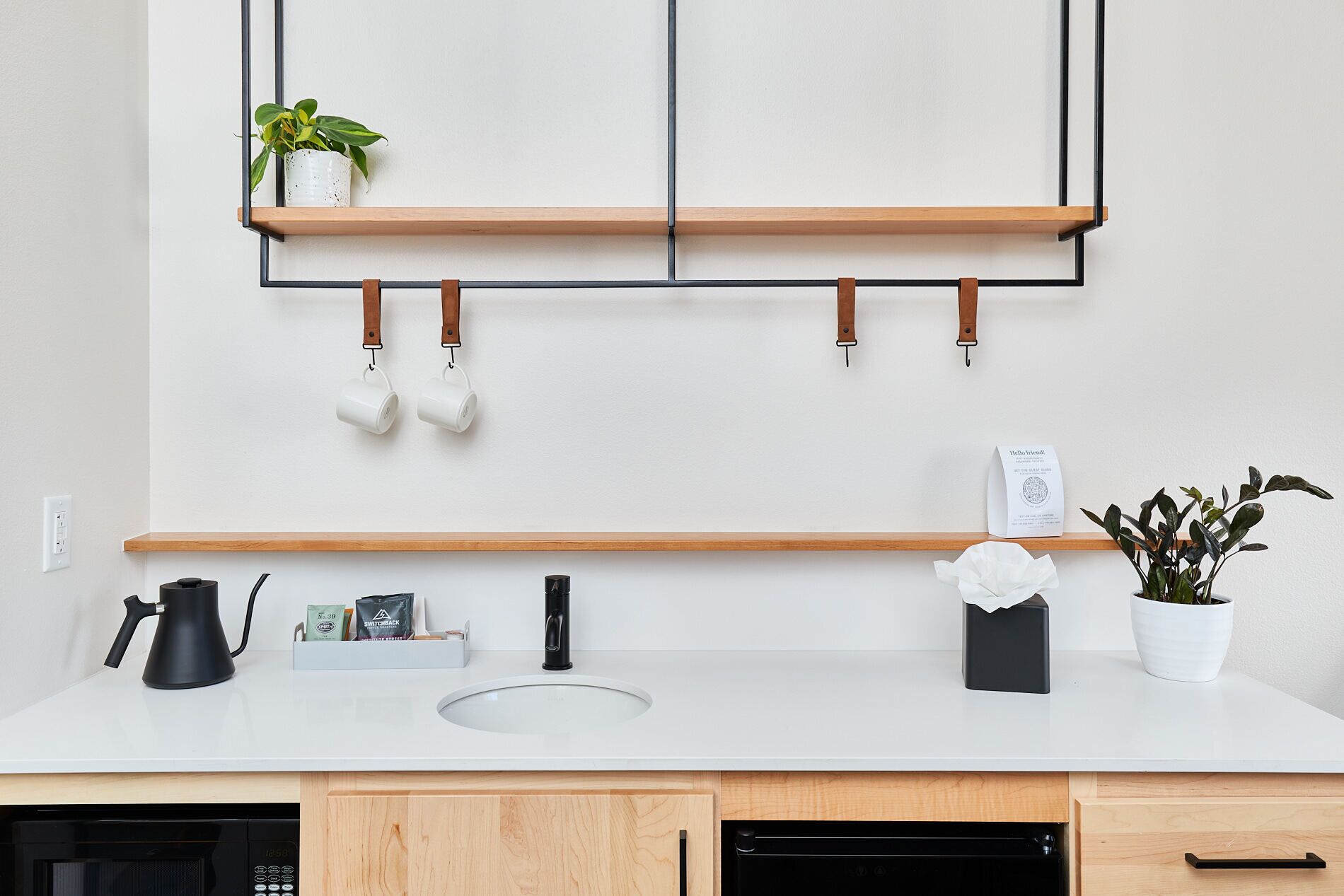 Une cuisine moderne avec des meubles en bois, un plan de travail blanc, un évier et une machine à café. On y voit deux tasses suspendues, une plante en pot et un petit panneau accroché au mur.