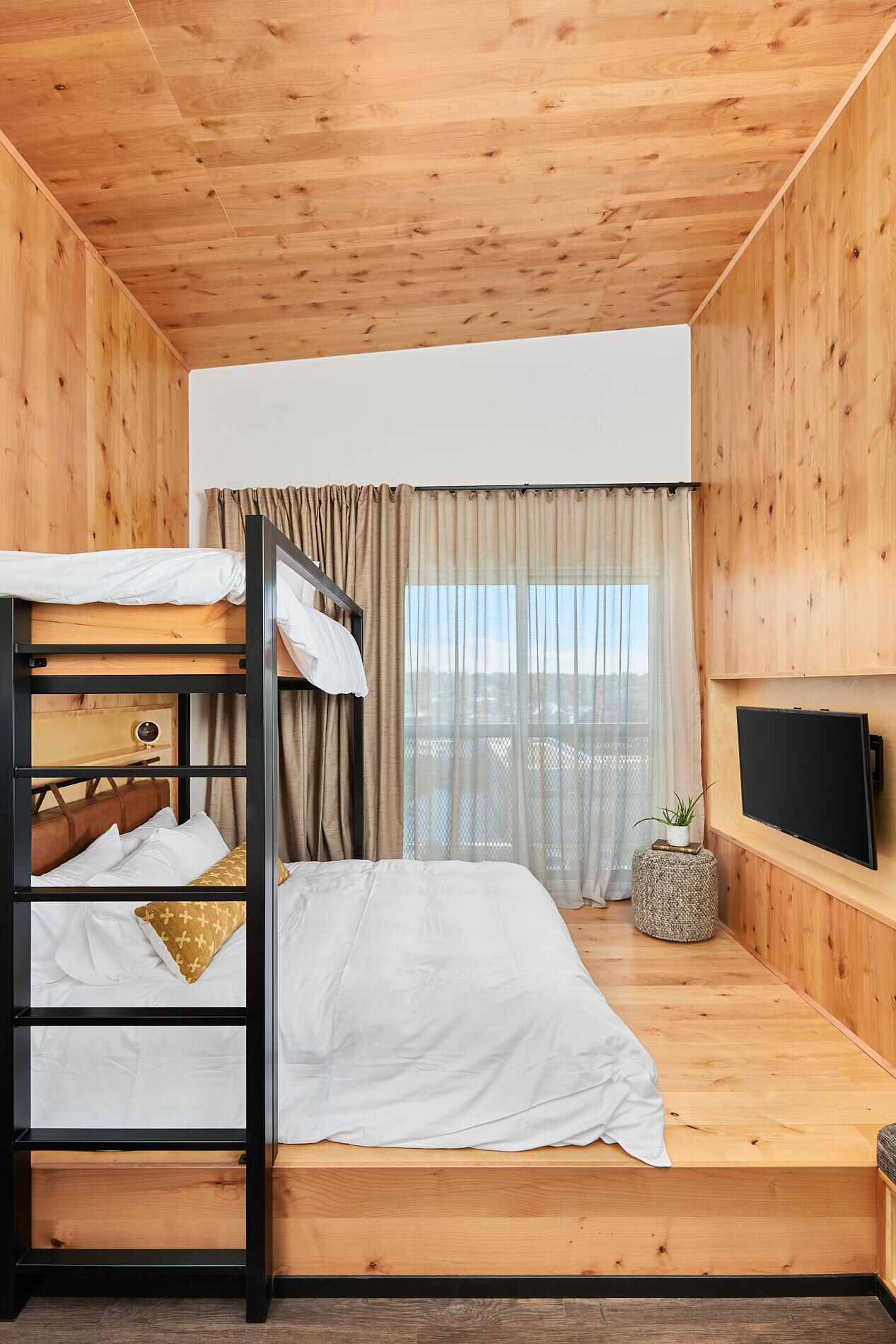 A bunk bed room with a wooden ceiling and walls, a television on the wall, and a window with curtains.