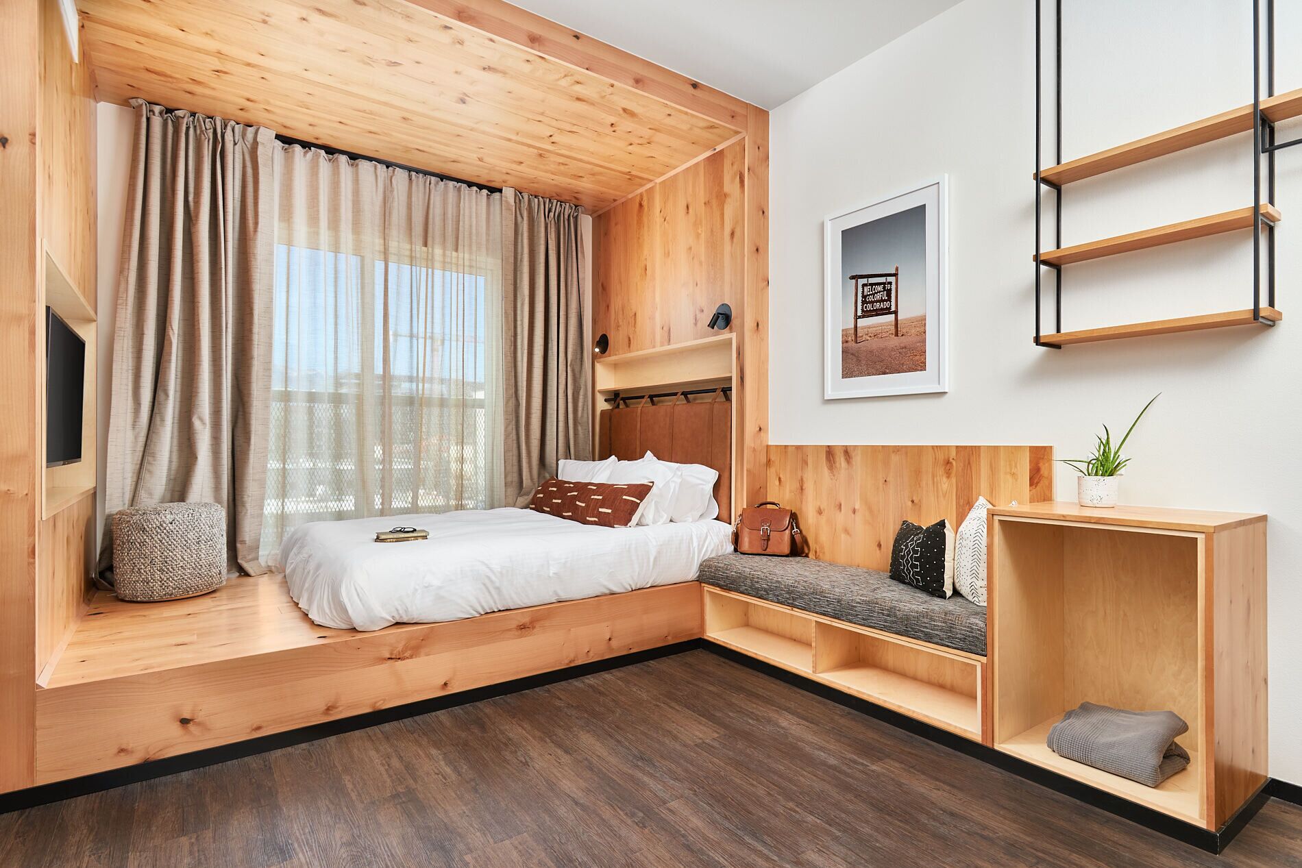 A modern bedroom with a wooden ceiling, a bed with white bedding, a bench with cushions, a small table, and a wall-mounted shelf.