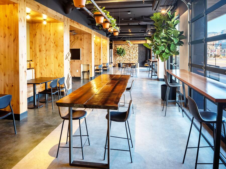 A spacious indoor dining area with long wooden tables and black chairs, large windows offering a view of the outdoors, and hanging plants adding greenery.