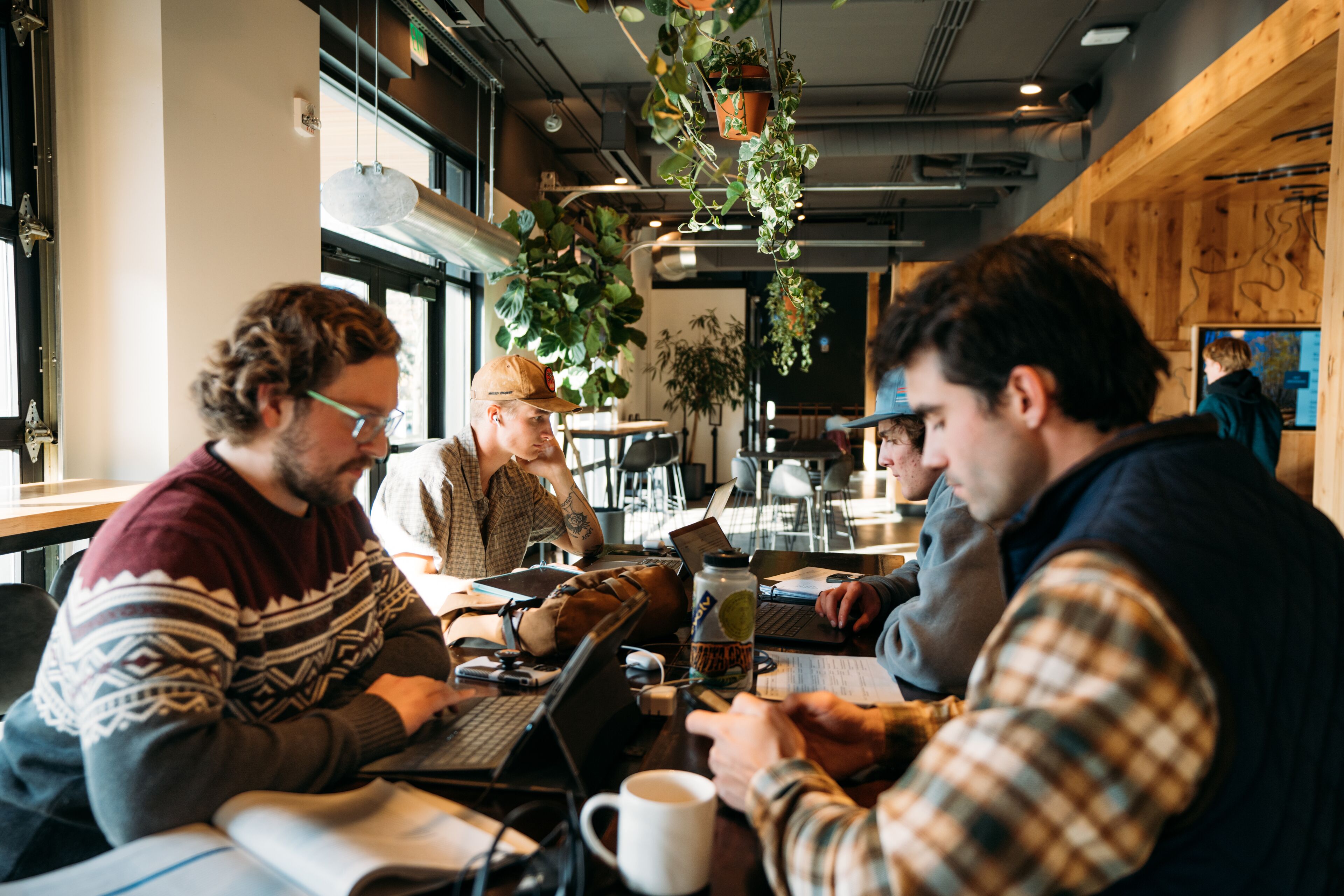 Un groupe de personnes est assis autour d’une table dans un café, devant des ordinateurs portables, avec une tasse de café et des plantes suspendues.