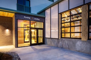 A building with a sign reading "Kinship Landing" and a large window with plants.