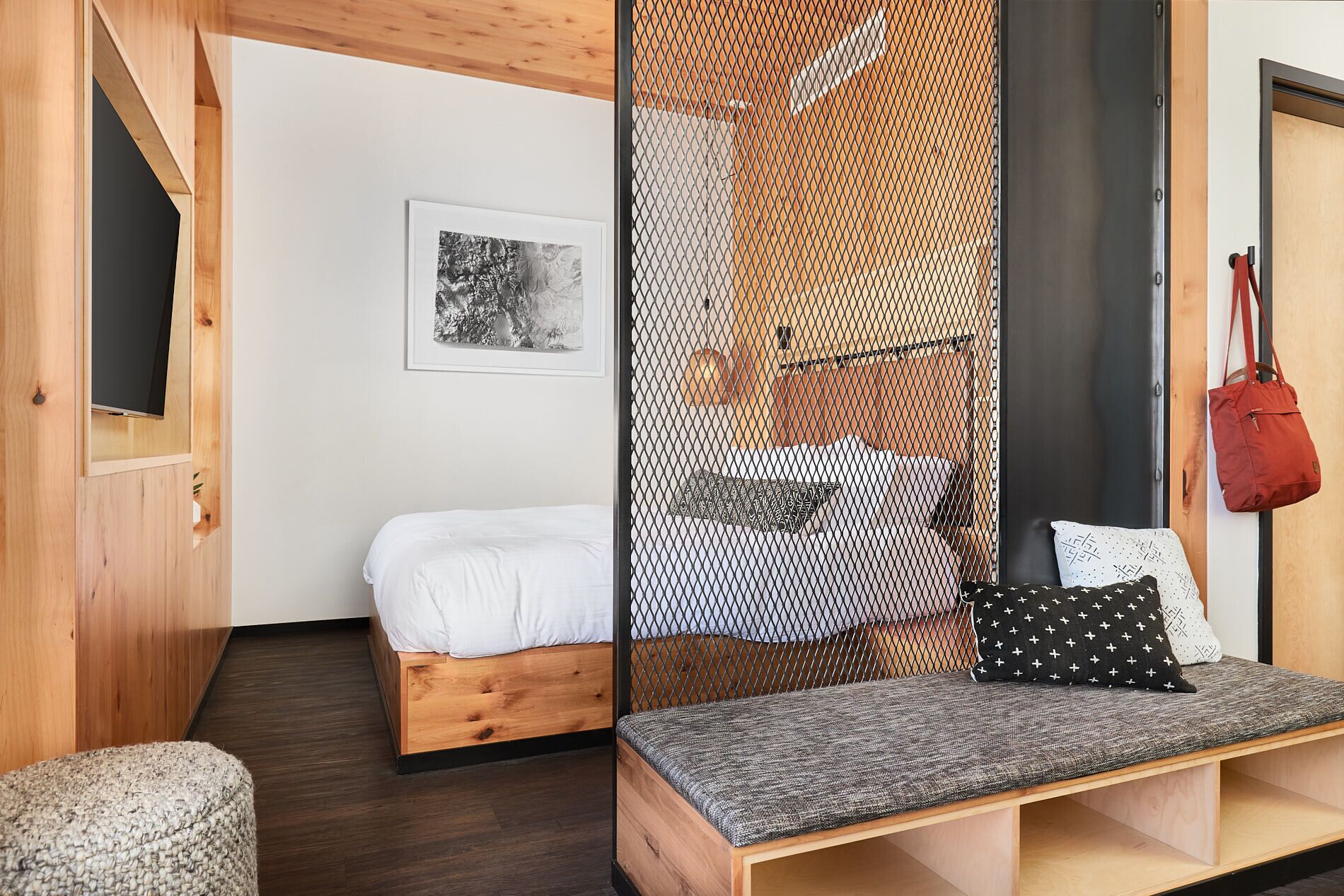 Una habitación moderna con una cama de madera, un banco con cojines, un cuadro enmarcado en la pared y un bolso rojo colgado en la puerta.