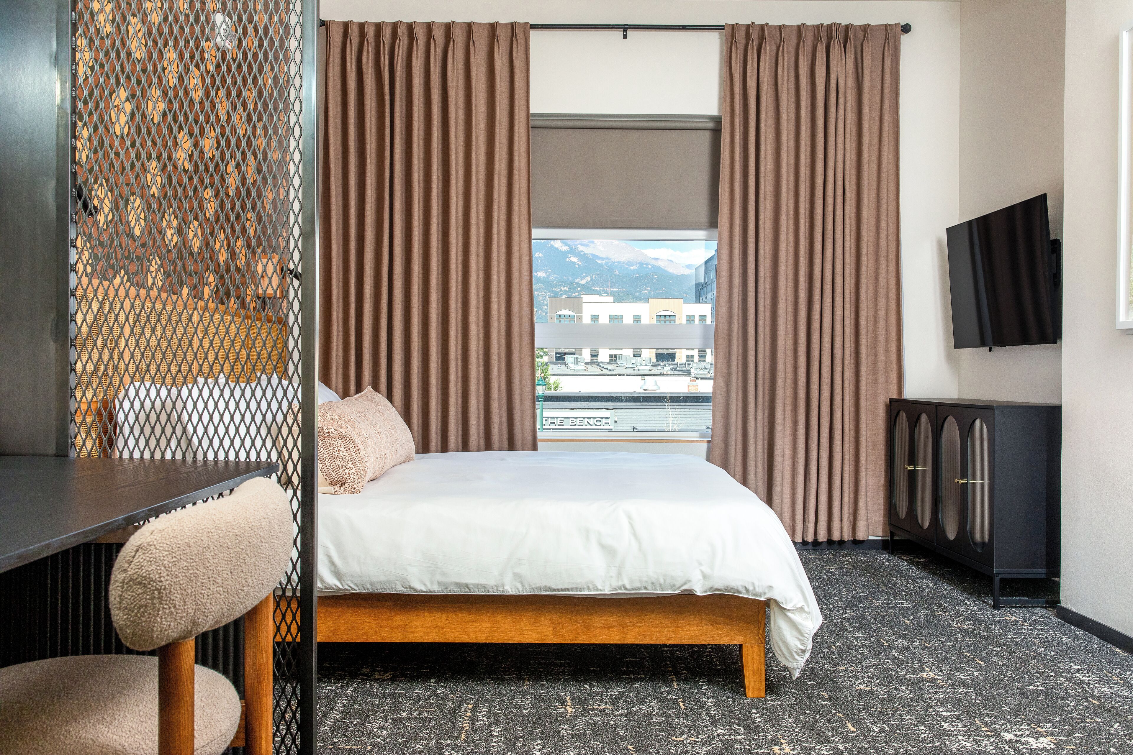 A hotel room with a bed, a desk, a chair, a TV, and a view of buildings and mountains.