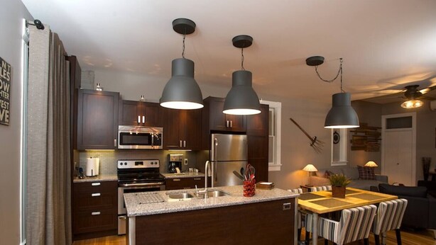 A modern kitchen with dark wood cabinets, stainless steel appliances, and a central island with a sink. There are three pendant lights above the island. - Vetehuset-Skorda Quiet Loft with Private Elevator (Lindsborg)