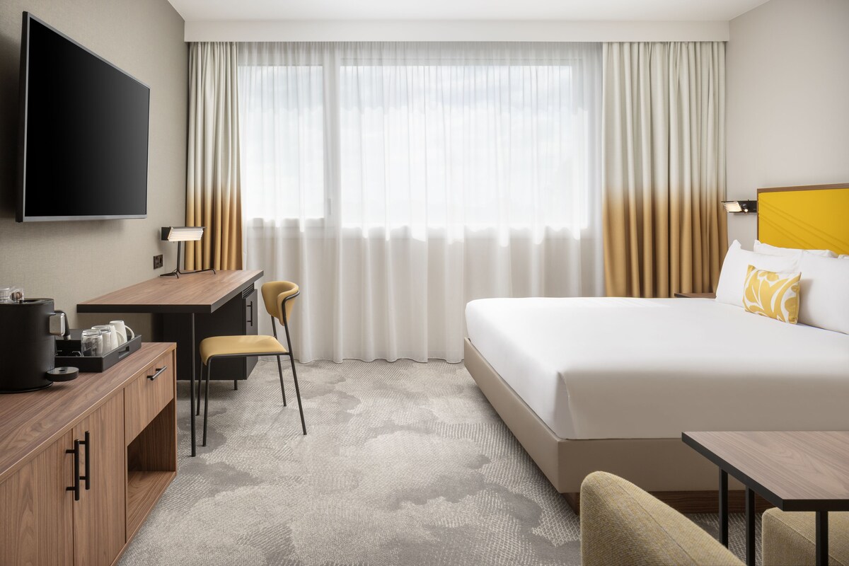 Habitación de hotel con cama, escritorio, silla, televisión y ventana con cortinas.