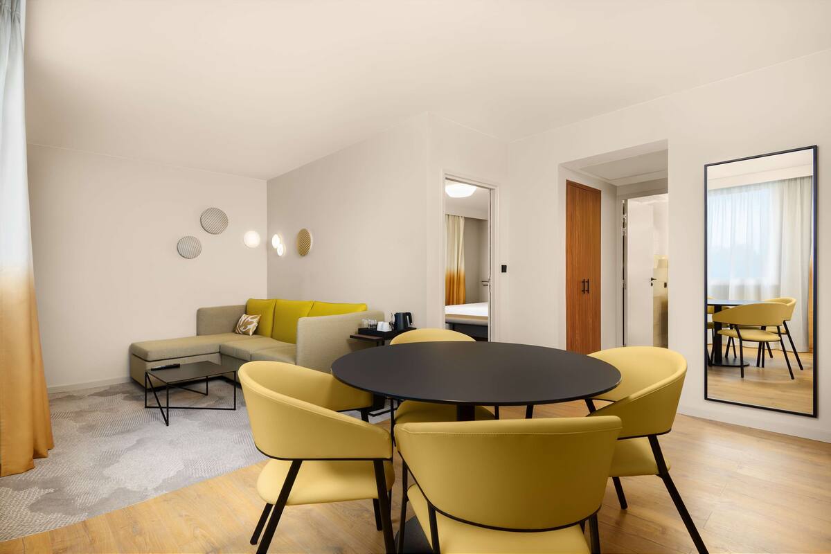 A modern living room with a sofa, yellow chairs, a round table, and a mirror reflecting the room.