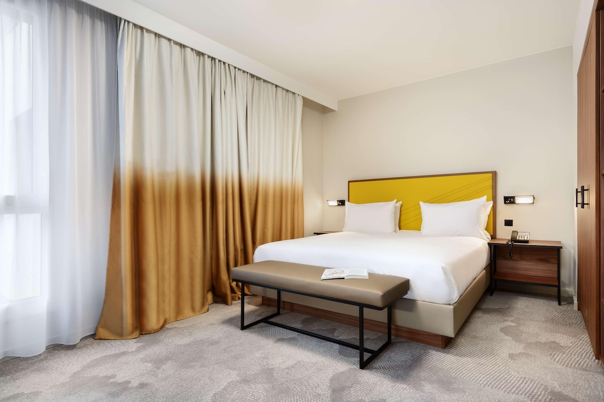 Una habitación de hotel con una cama grande, una mesita de noche, un banco y un ventanal con cortinas.