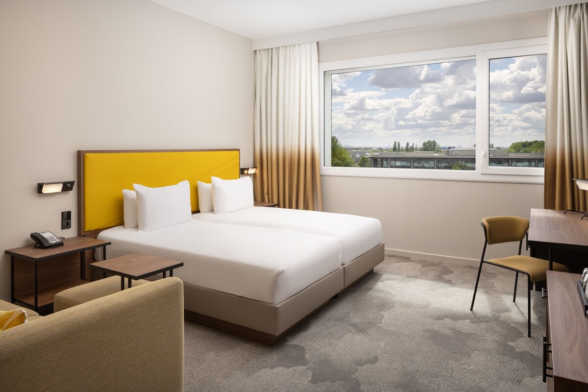 Habitación de hotel con cama, mesita de noche, silla, escritorio y vistas a la ciudad desde la ventana.