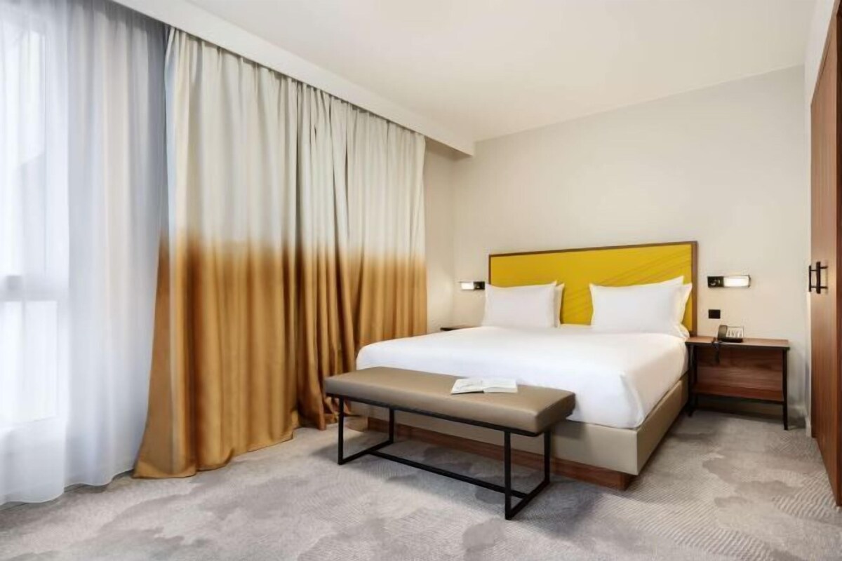 Una habitación de hotel con una cama, un banco, una mesita de noche y cortinas.