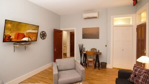 Un salon avec un canapé gris, un téléviseur fixé au mur, un coin repas avec une table et des chaises, et une porte blanche.