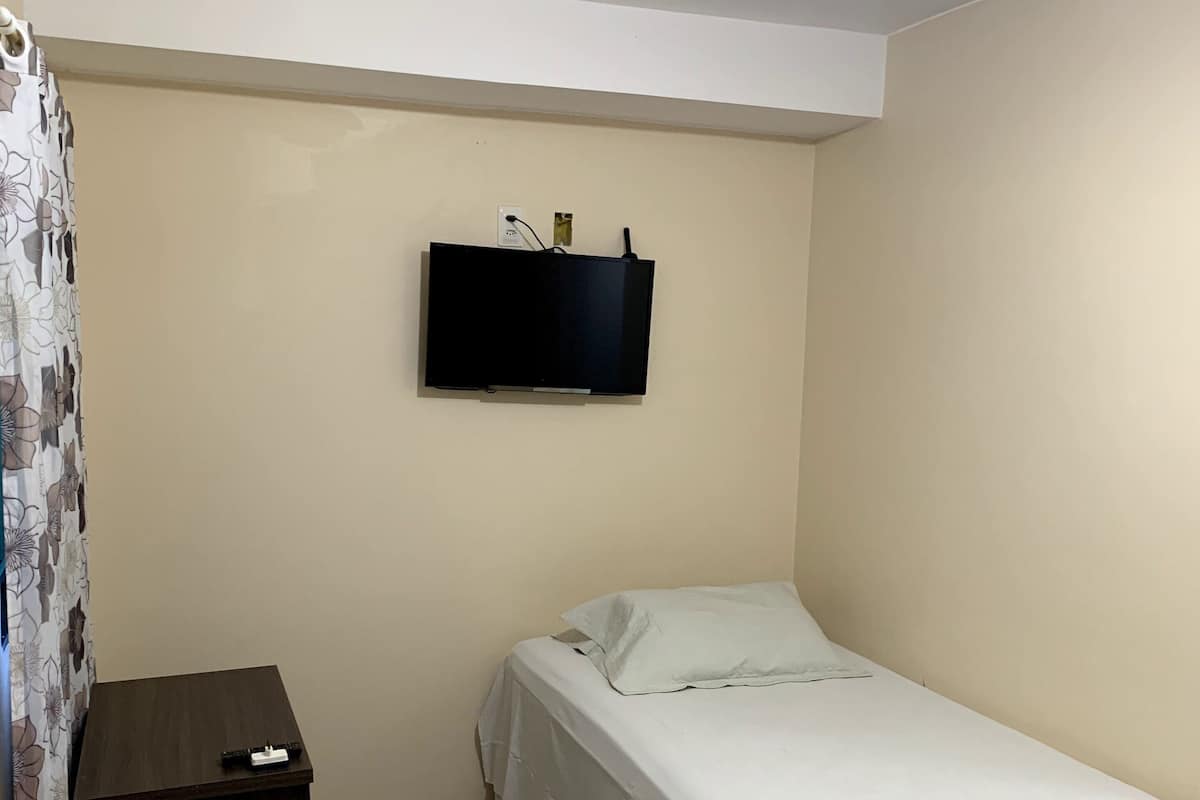 Una habitación pequeña y limpia con una cama, un televisor montado en la pared y una mesita con mantel blanco.
