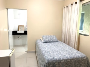 Un dormitorio con cama, una mesita de noche, ventana con cortinas y baño con lavamanos y espejo.