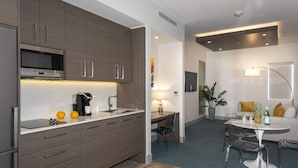 Una cocina moderna con microondas, cafetera y naranjas sobre la encimera. Hay un comedor con mesa y sillas, y un sofá con una planta y una botella.