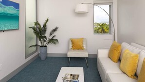 Una sala de estar moderna con un sofá blanco, cojines amarillos, un sillón blanco, una planta grande en maceta, una mesa de centro con revistas, un televisor montado en la pared y una ventana con vistas a palmeras.