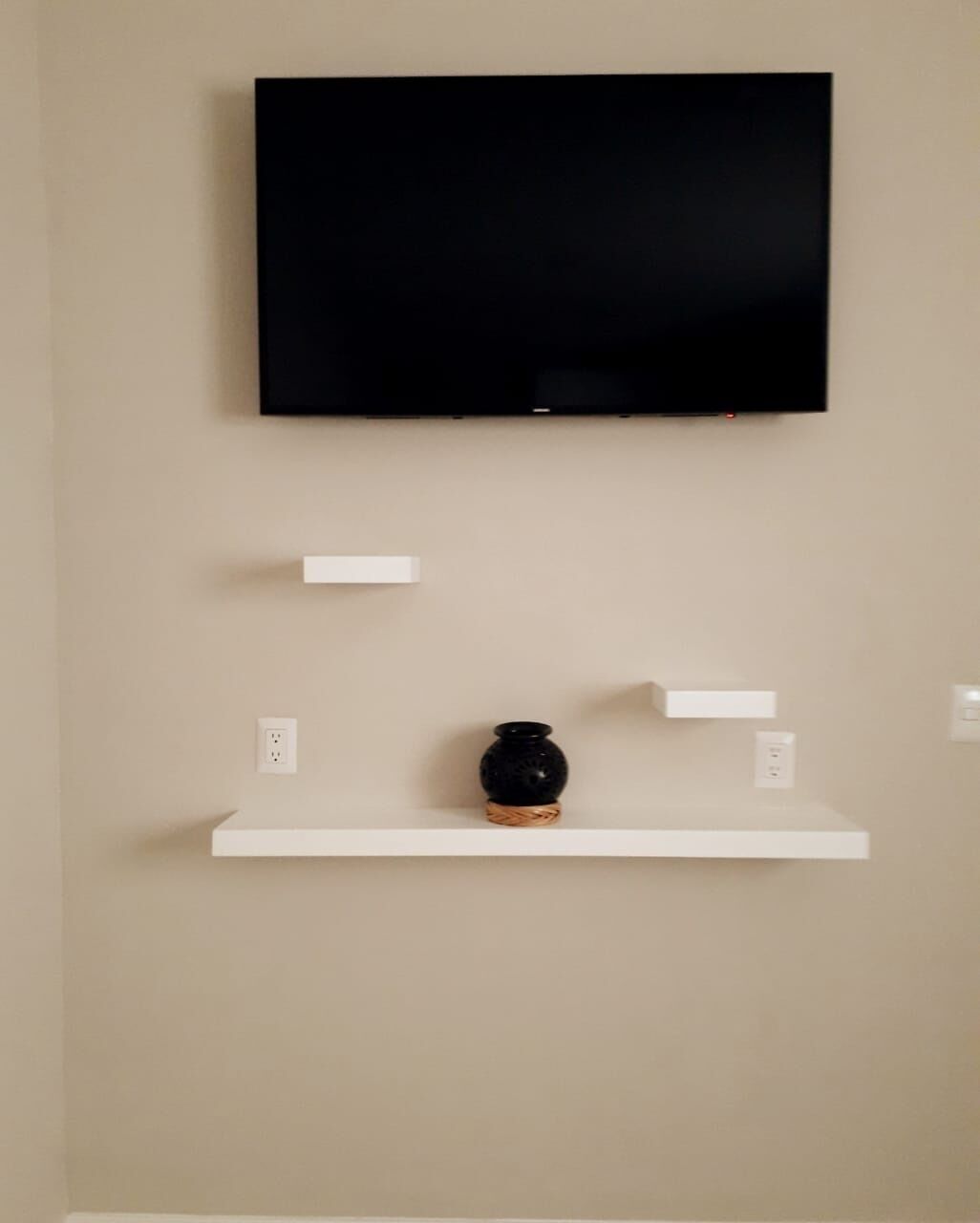 Un téléviseur fixé au mur, au-dessus d’une étagère blanche, avec un vase noir.