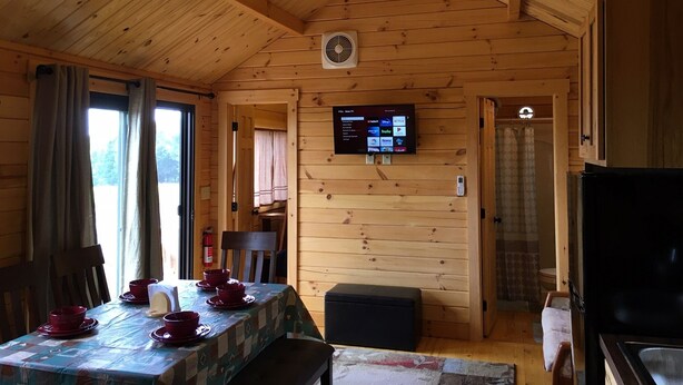 Een houten blokhutinterieur met een eettafel voor vier personen, een plafondventilator en een aan de muur bevestigde tv waarop streamingdiensten worden weergegeven.