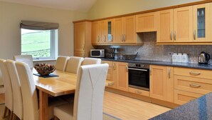 A modern kitchen with wooden cabinets, a microwave, and a stove. There is a dining table with chairs and a view of a green field through a window.
