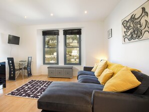 A modern living room with a grey sofa, a patterned rug, a small table, and two windows with black blinds. - 1 bedroom accommodation in Looe (Looe)