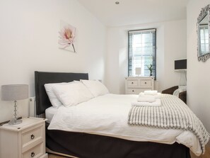 A neatly arranged bedroom with a bed, bedside table, lamp, and a window with blinds. - 1 bedroom accommodation in Looe (Looe)