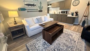 A living room with a white sofa, a wooden coffee table, a patterned rug, a kitchen area with cabinets and appliances, and a wall with nautical decor. - Lakeside Serenity: Charming One-Bedroom Villa (Bridgman)