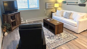 A living room with a TV, sofa, coffee table, and decorative items including a wall-mounted sign reading "WEKO BEACH". - Lakeside Serenity: Charming One-Bedroom Villa (Bridgman)