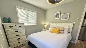 A bedroom with a bed, dresser, window, artwork, and a refrigerator. - Lakeside Serenity: Charming One-Bedroom Villa (Bridgman)