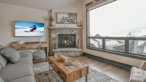 A living room with a stone fireplace, a large TV, and a view of a snowy landscape through a large window. - Alders 46 Outpost Dr (Keystone)