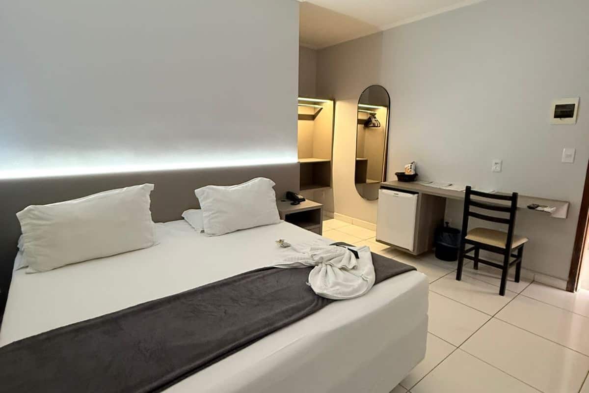 A hotel room with a large bed, a desk, a chair, a mirror, and a small kitchenette.