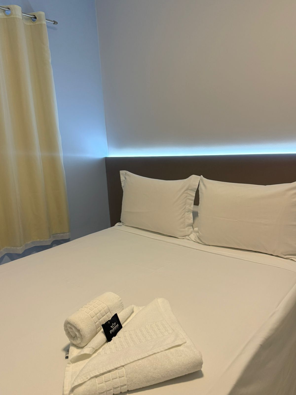 Ein Hotelzimmer mit einem ordentlich bezogenem Bett, einem Kopfkissen, einem gefalteten Handtuch und einem Vorhang.