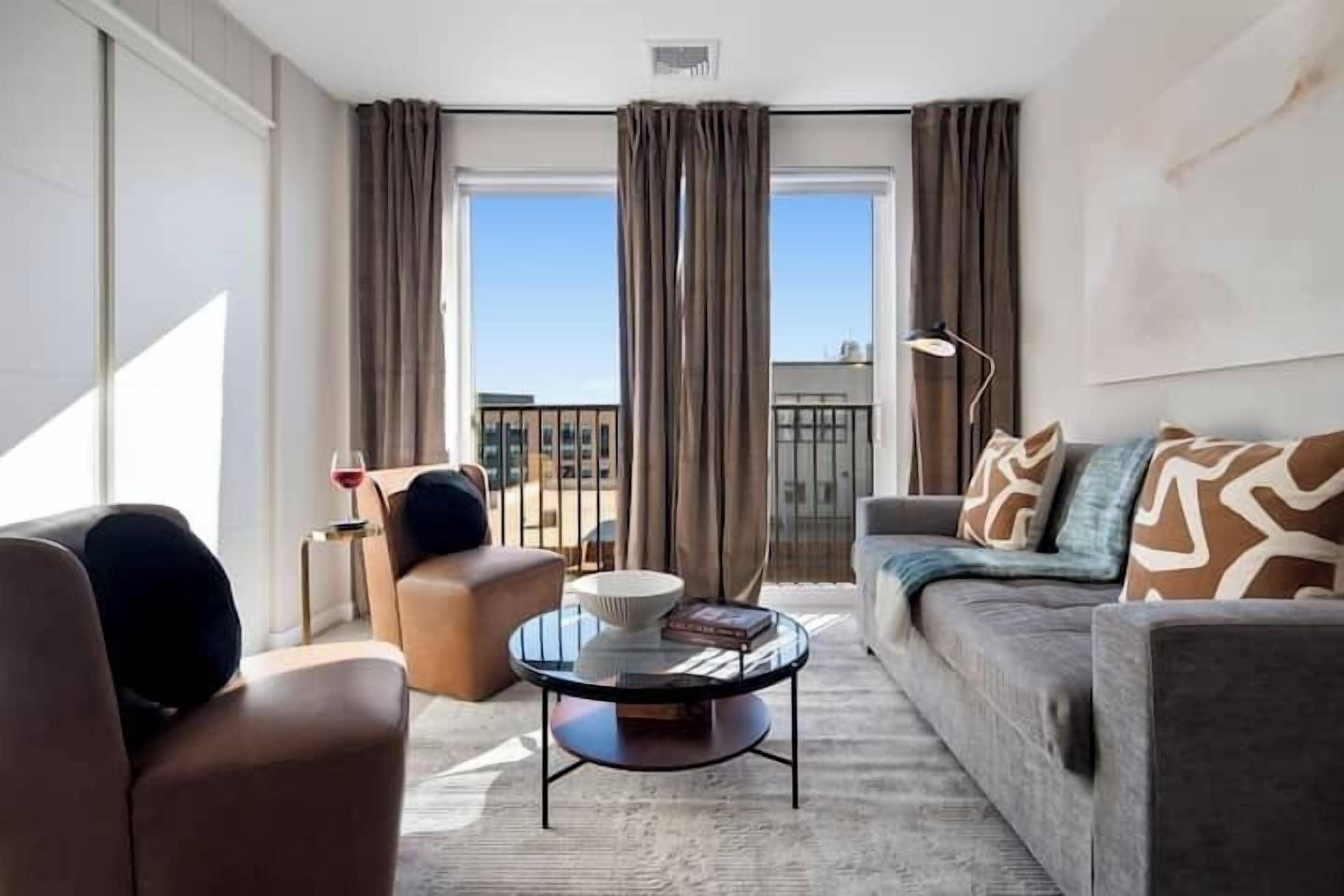 A modern living room with a sofa, armchair, and coffee table. There is a balcony with curtains and a view of buildings.