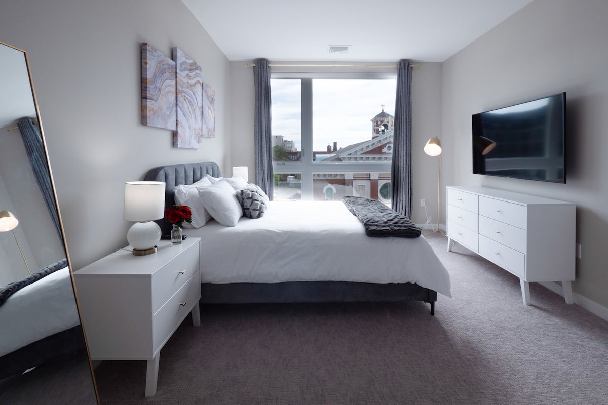 A modern bedroom with a large bed, a TV, a dresser, and a window with a city view.