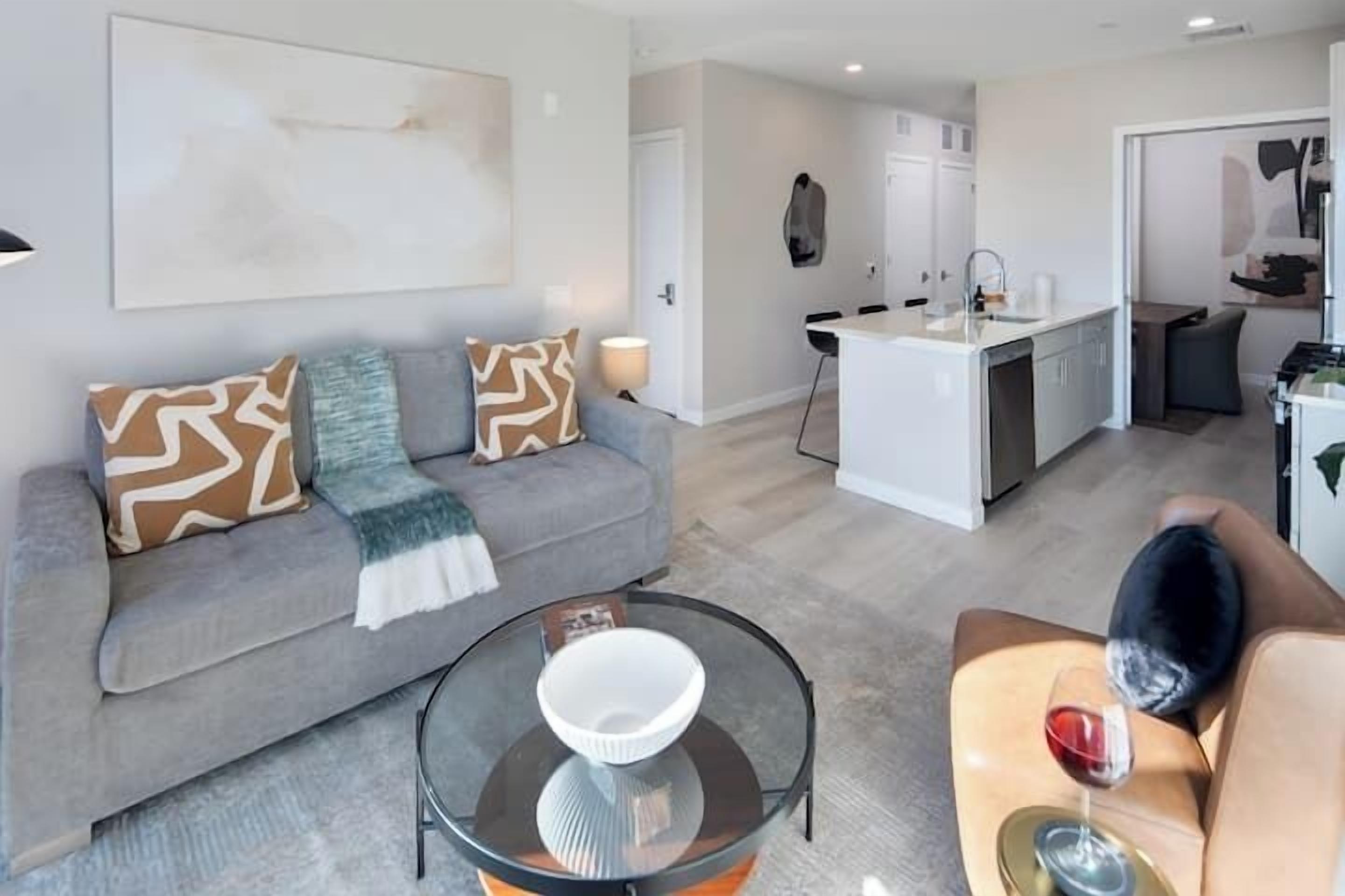 Una sala de estar moderna con un sofá gris, una mesa de centro de vidrio y un comedor con una cocina blanca al fondo.
